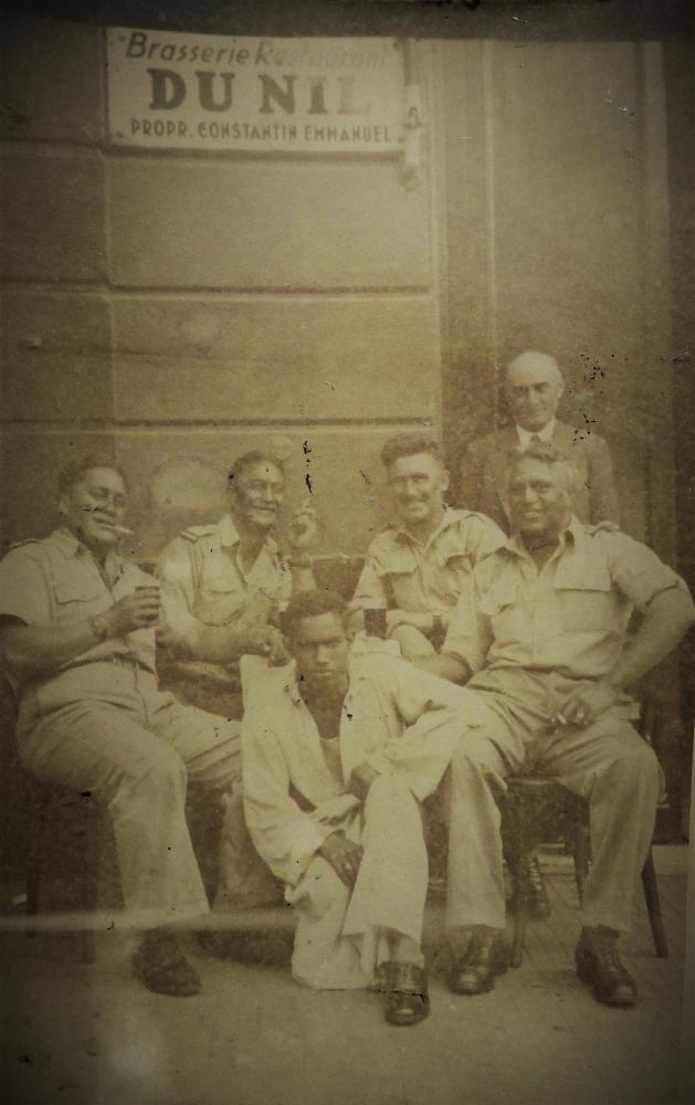 Four New Zealand soldier sit smoking while a man sits on the ground in front of them. An older man stands behind them.