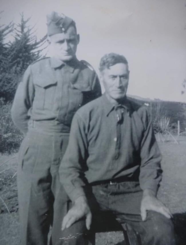 A solider in uniform stands behind a seated older man.