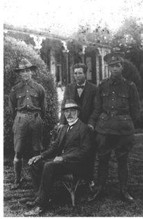 Four men in front of a house. Three men are standing, two in uniform and one in a suit, while the fourth, much older man, is seated. 