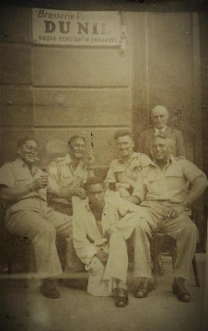 Four New Zealand soldier sit smoking while a man sits on the ground in front of them. An older man stands behind them.